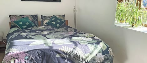 A bed with a floral bedspread, two pillows, a framed picture above the bed, and a window with a plant.