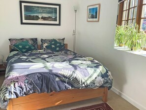 A bed with a floral bedspread, two pillows, a framed picture above the bed, and a window with a plant. - This cosy cottage is an ideal base for you and your dogs. (St Just)