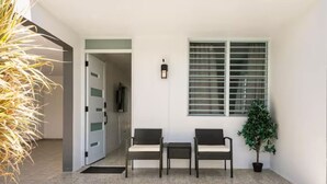 Uma entrada luminosa com porta, duas cadeiras, uma mesinha e um vaso de planta.