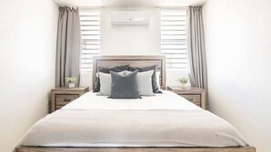 Quarto com uma cama grande, duas mesas de cabeceira, ar condicionado na parede e cortinas nas janelas.