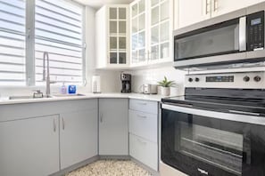Una cocina moderna con armarios blancos, microondas, horno, cafetera y una ventana con persianas.