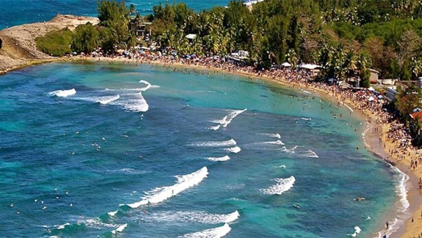 Uma praia costeira com águas cristalinas, areal arenoso e área densamente povoada com edifícios e árvores.