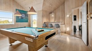 Een biljart tafel in een ruimte met houten wanden en plafond, een dartbord, en een bar met stoelen.