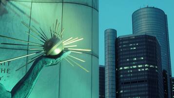 A modern sculpture with a spherical element and radiating lines, set against a backdrop of tall buildings with reflective glass facades.