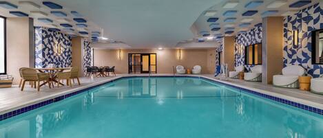 A large indoor swimming pool with tiled walls and ceiling, surrounded by lounge chairs and tables.