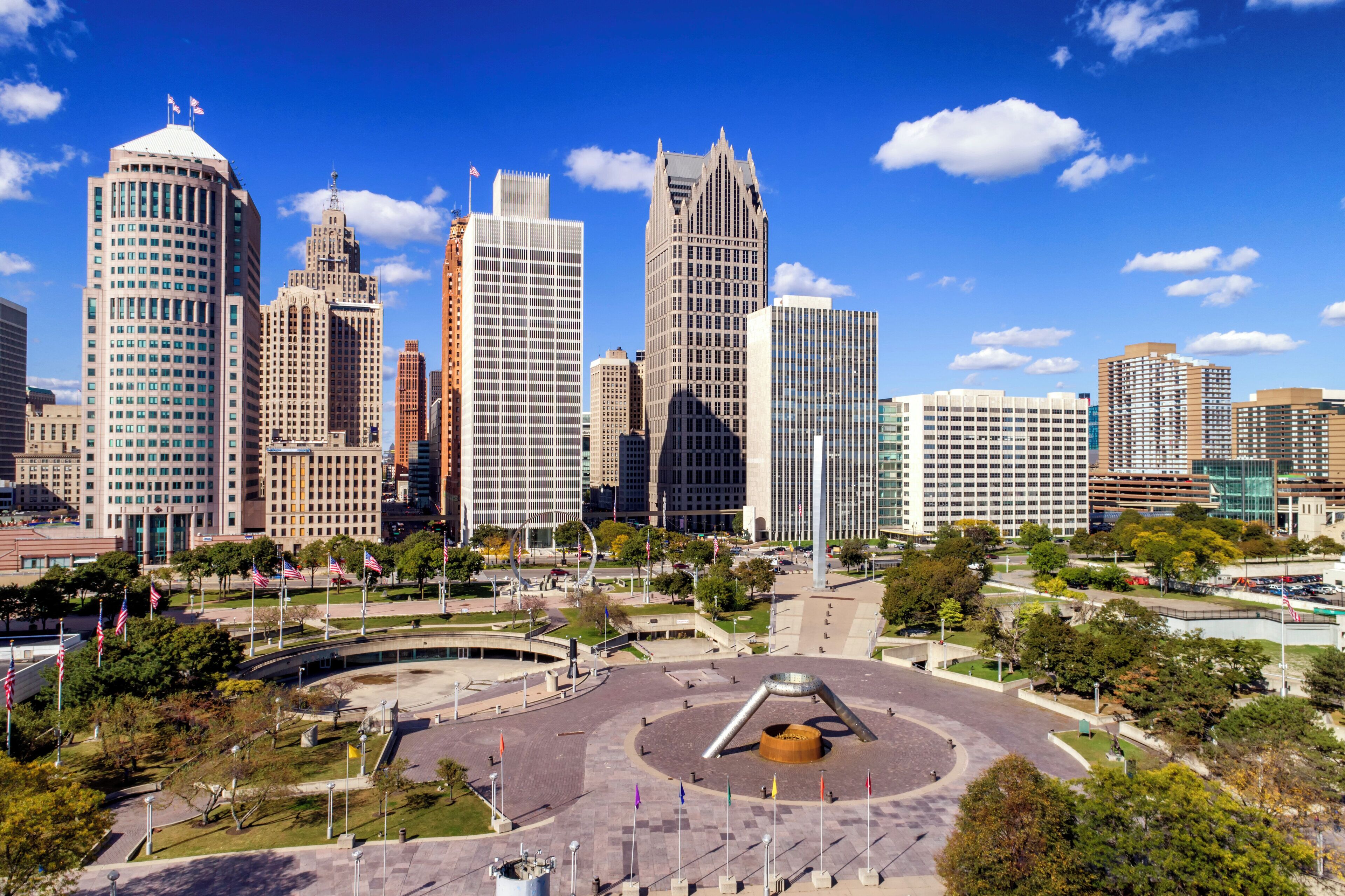 A cityscape with modern skyscrapers, a central plaza with a circular monument, and a clear blue sky.