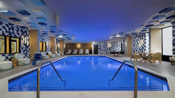 An indoor swimming pool with a blue tiled floor, surrounded by lounge chairs and a seating area with a patterned wall. - EVEN Hotel Detroit North - Troy (Troy)