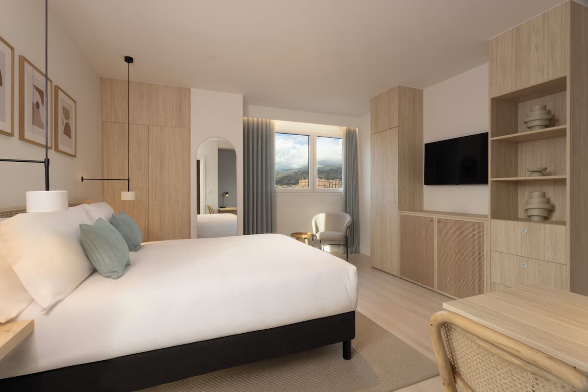 A modern hotel room with a large bed, a TV, a desk, and a view of the city.