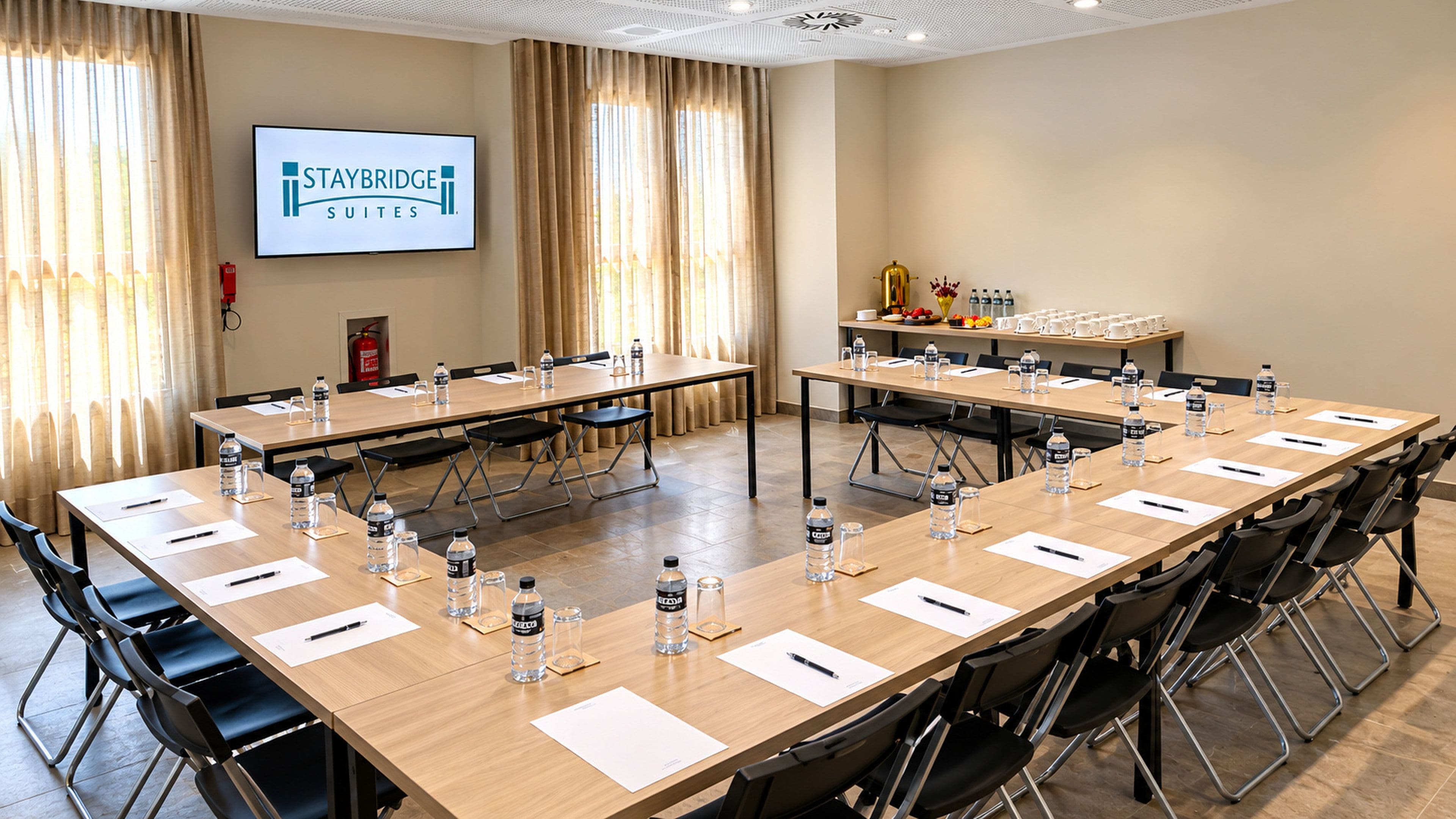 Una sala conferenze con la marca Staybridge Suites, dotata di tavoli, sedie, bottiglie d'acqua e uno schermo per proiettore.