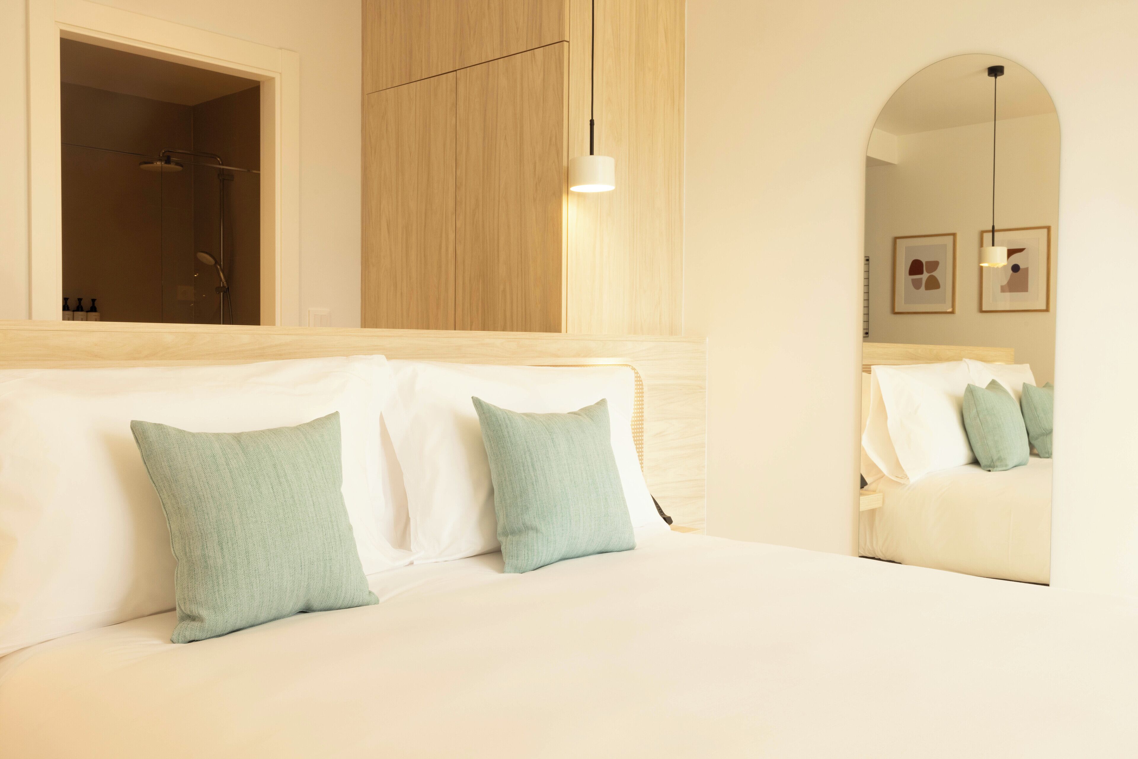 A hotel room with a wooden headboard, a bed with white linens and green pillows, a shower, and a hanging light fixture.