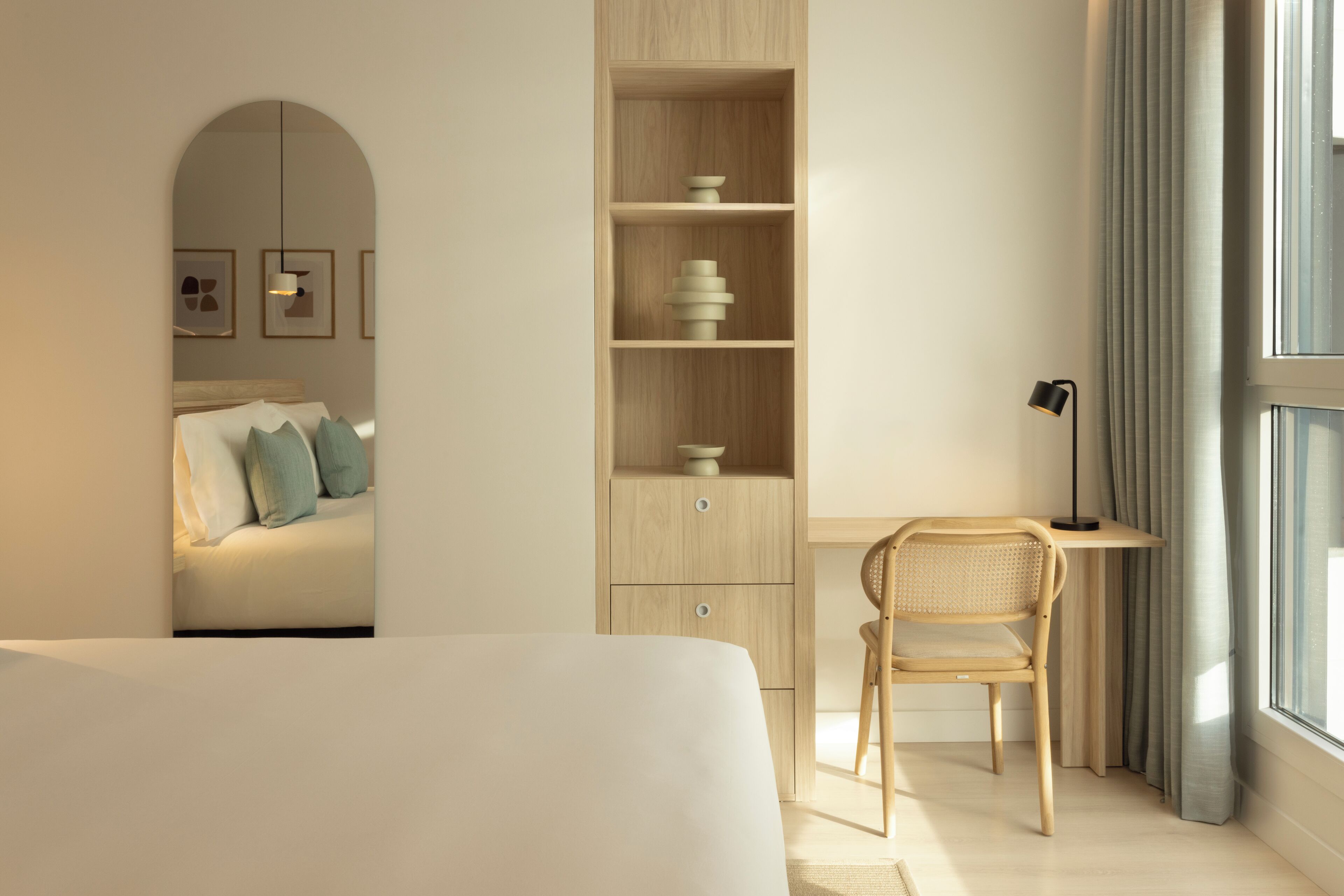 A modern bedroom with a bed, a desk, and a chair. There is a bookshelf with decorative items and a lamp on the desk.