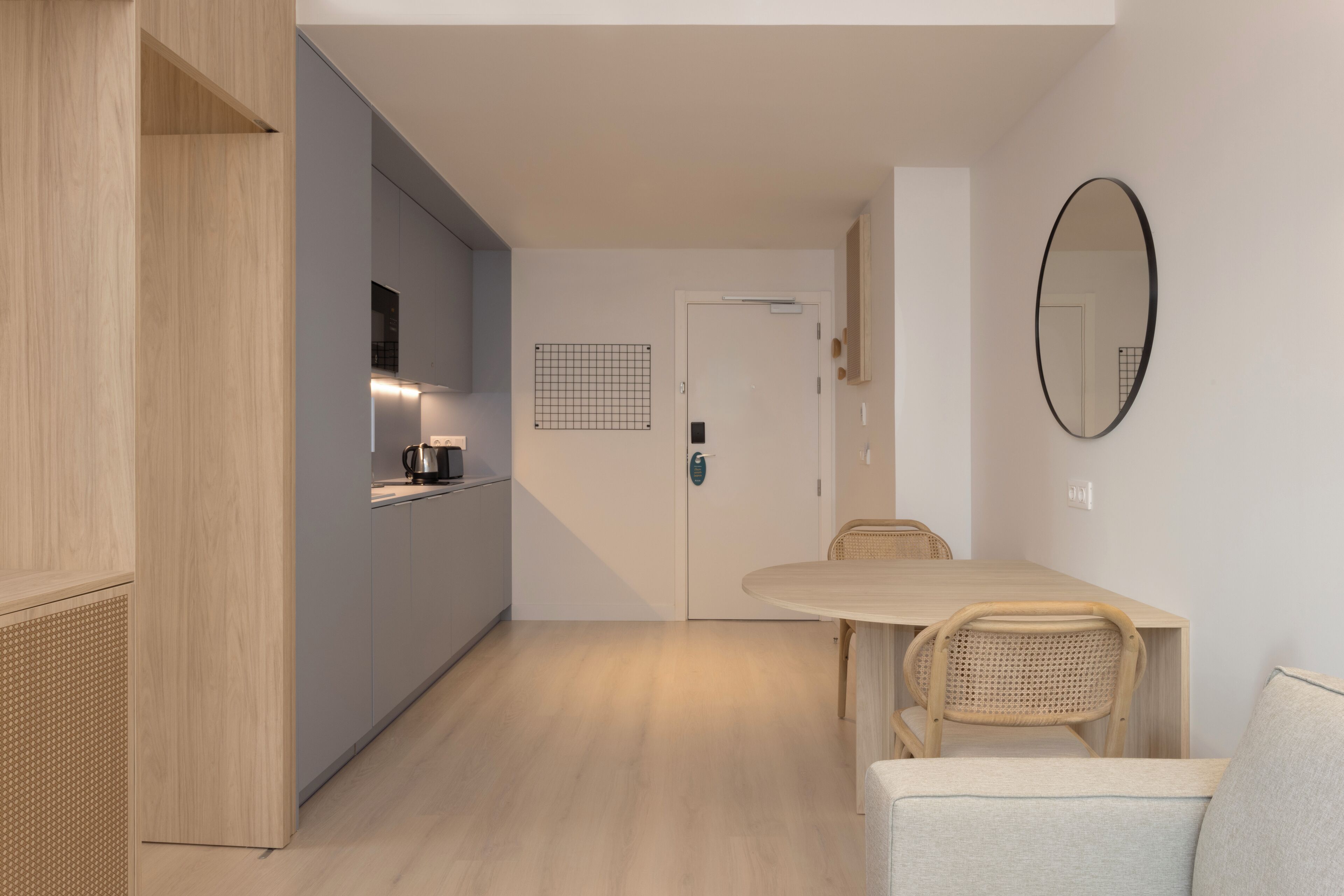 Una cocina moderna con estufa integrada, una zona de comedor con una mesa redonda y sillas de mimbre, y una puerta blanca con decoración mural de diseño de cuadrícula.