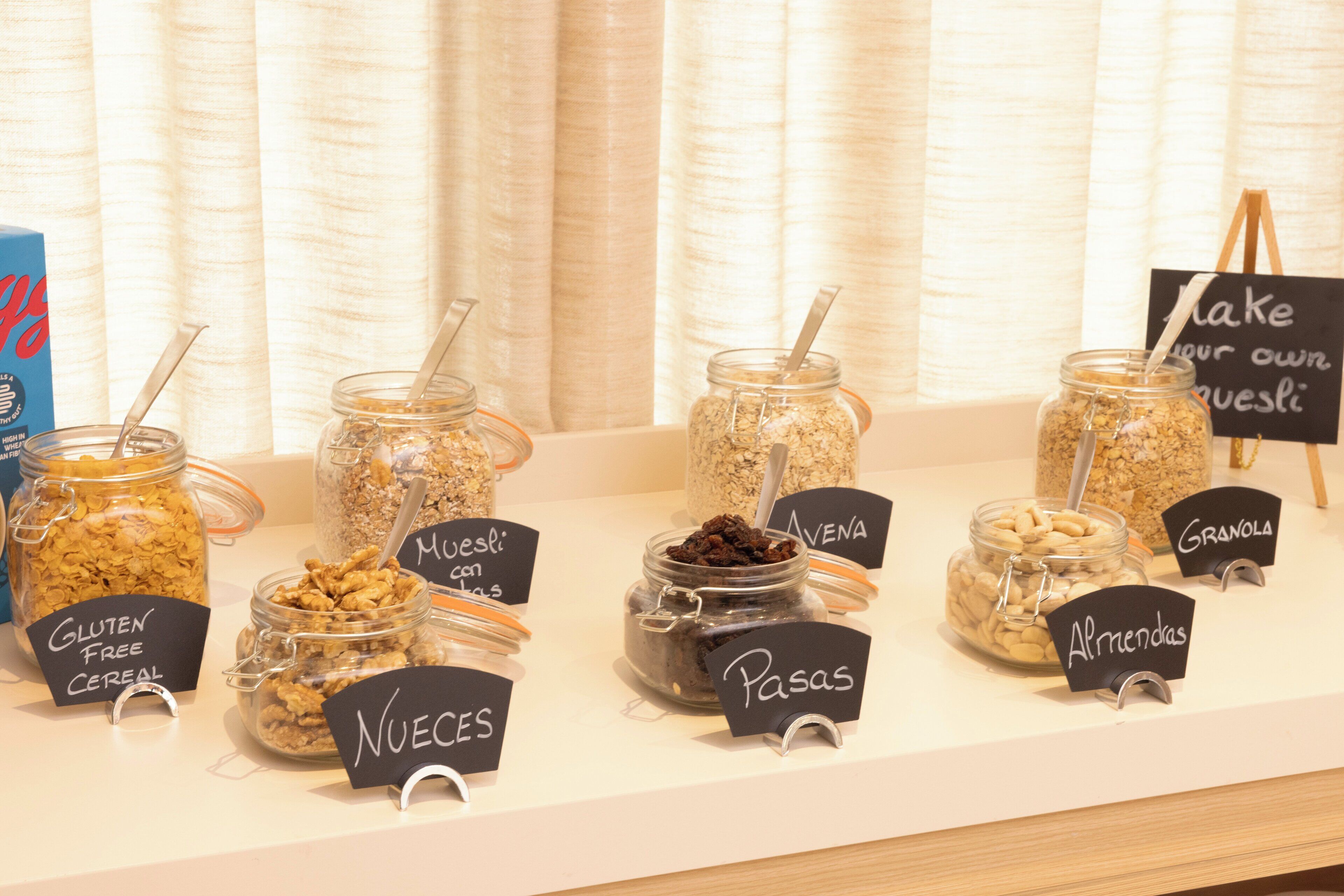A variety of food items in jars labeled with names like "Gluten Free Cereal," "Nueces," "Muesli con Avena," "Pasas," and "Almendras" on a counter.