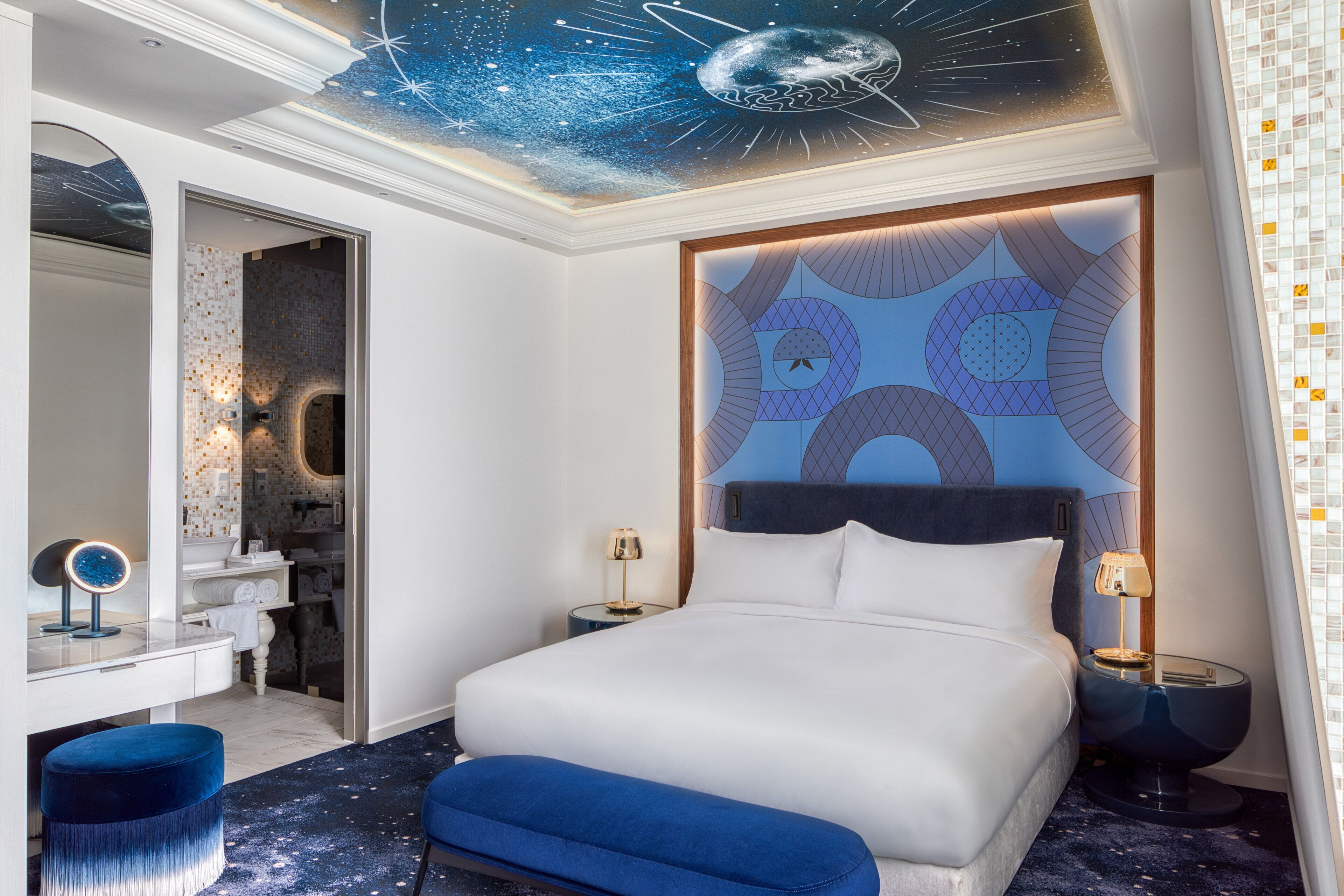 A modern hotel room with a large bed, a vanity with a mirror, and a ceiling with a space-themed design.