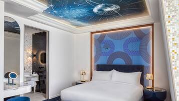 A modern hotel room with a large bed, a vanity with a mirror, and a ceiling with a space-themed design.