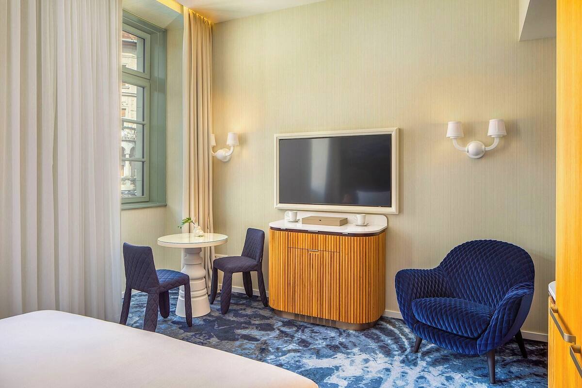 Ett hotellrum med en säng, en tv, två stolar, ett litet bord och ett fönster med gardiner.