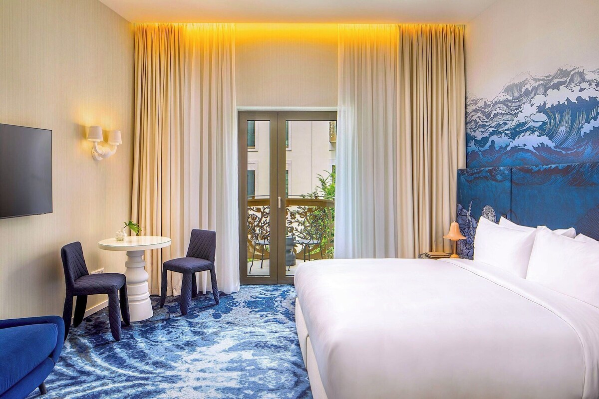 Een hotelkamer met een bed, een bureau met twee stoelen, een televisie en een groot schilderij aan de muur.