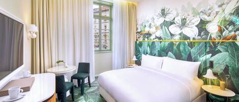 Habitación de hotel con cama, escritorio con silla, una pequeña mesa con otra silla y un amplio ventanal con cortinas.