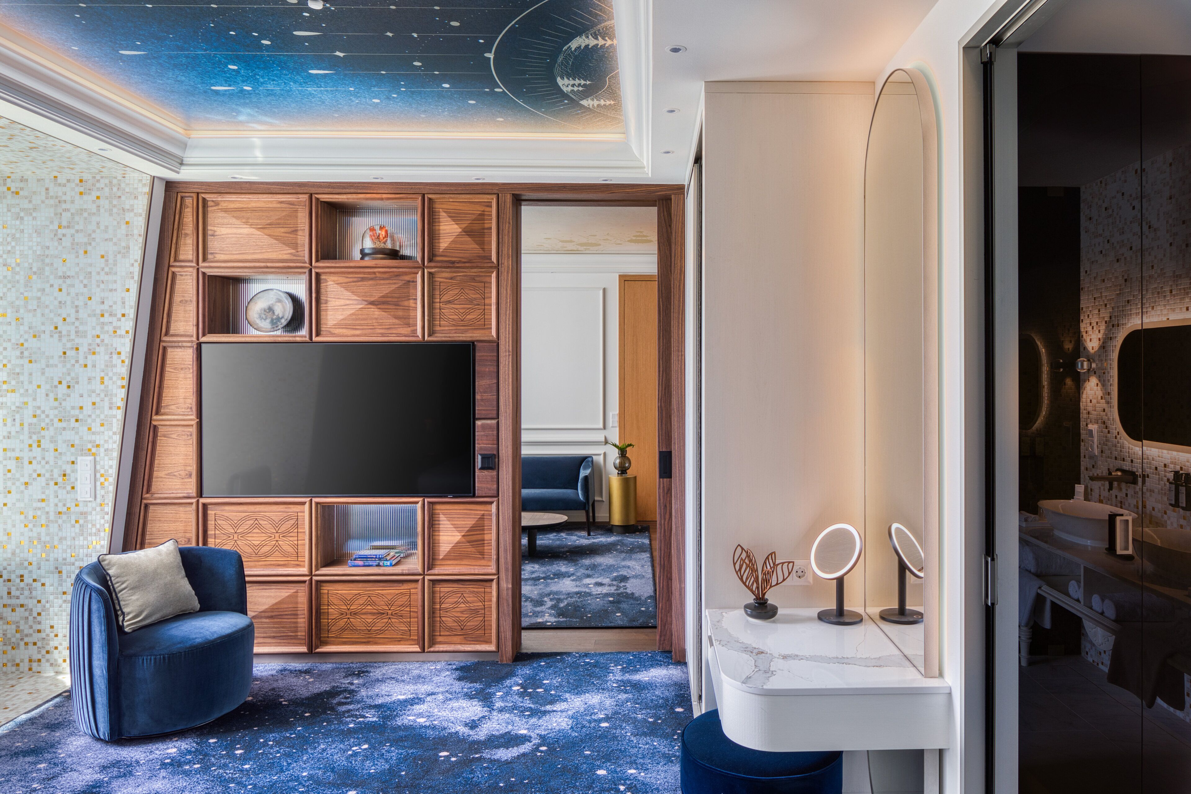 A modern room with a starry ceiling, a large flat-screen TV, a blue sofa, a white marble sink, and a bathroom with a mirror.