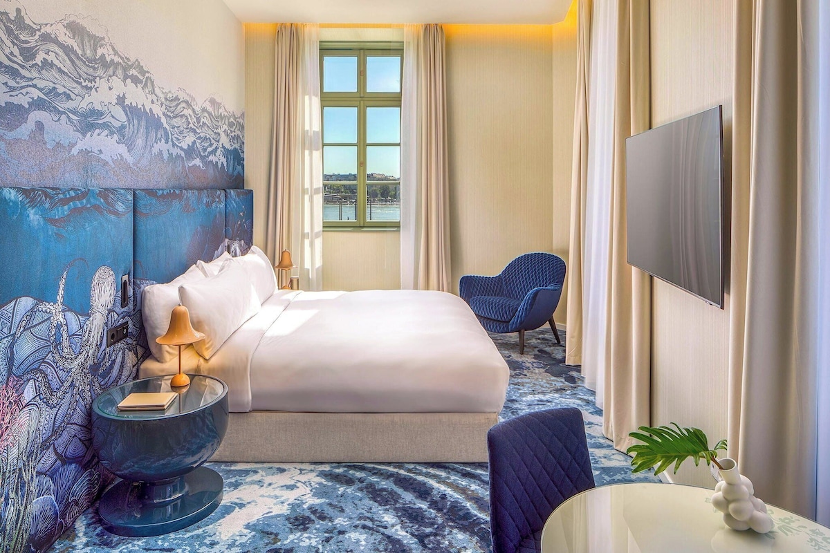 Een hotelkamer met een bed, een glazen bijzettafeltje, een stoel, een televisie en uitzicht op het groen buiten.