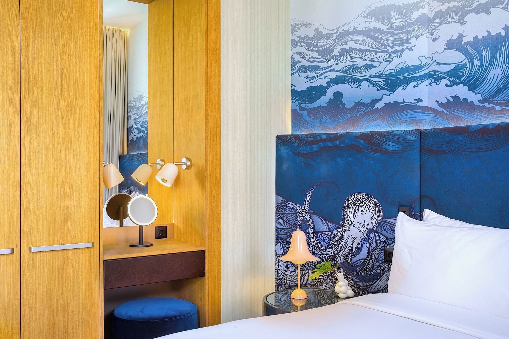 A hotel room with a bed, a bedside table with a lamp, a mirror, and a wall with a wave painting.