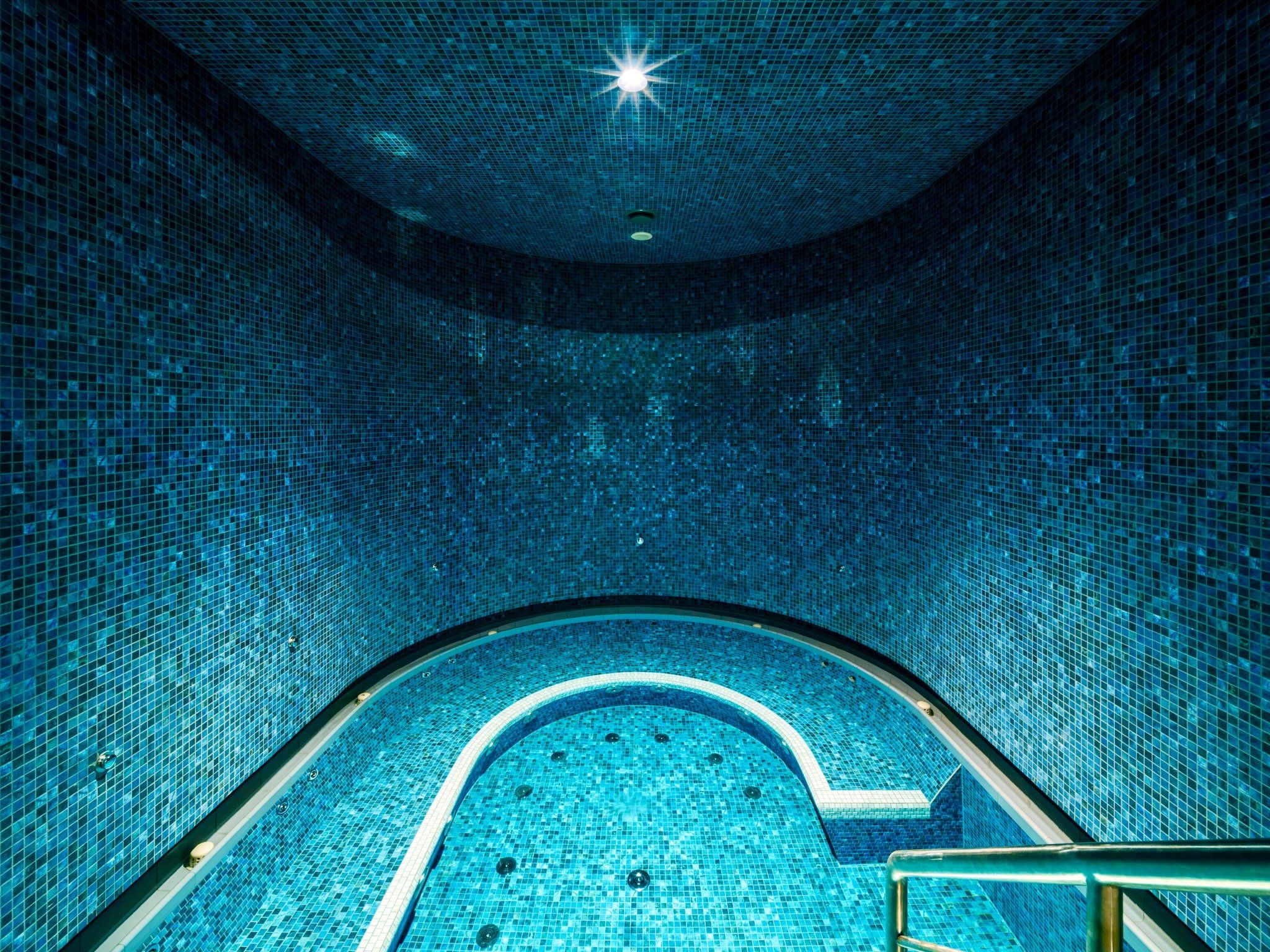 An indoor swimming pool with a tiled floor and walls, a curved edge, and a central light fixture.