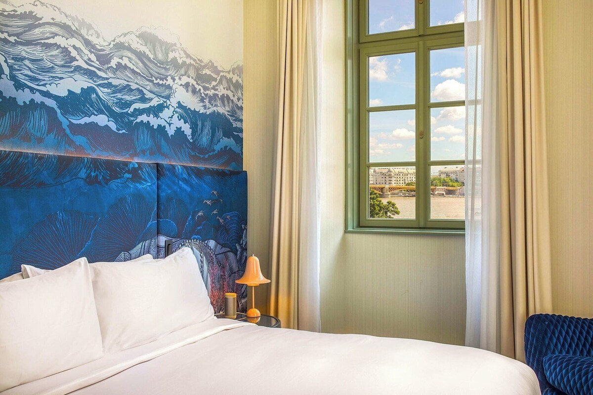 Een hotelkamer met een bed, een nachtkastje met lamp, een raam met gordijnen en een muurschildering.