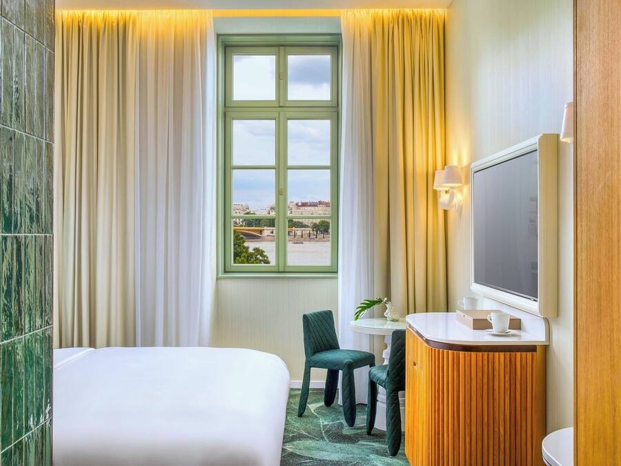 Habitación de hotel con cama, escritorio con silla, baño y ventana con vista a un puente y edificios.
