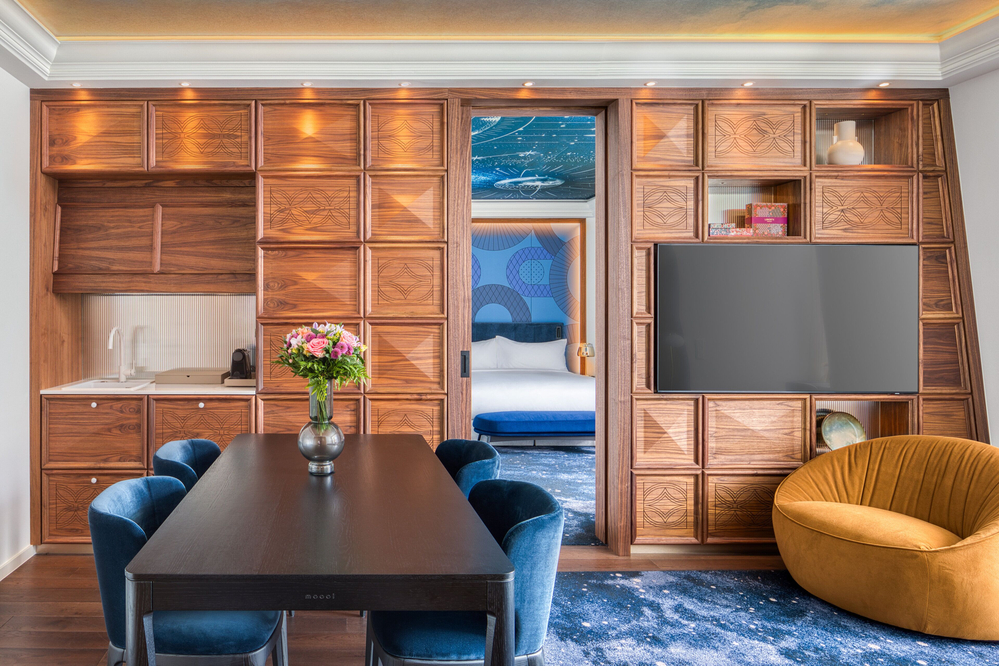 A modern living room with a wooden panel wall, a dining table with chairs, a sofa, a TV, and a decorative vase.