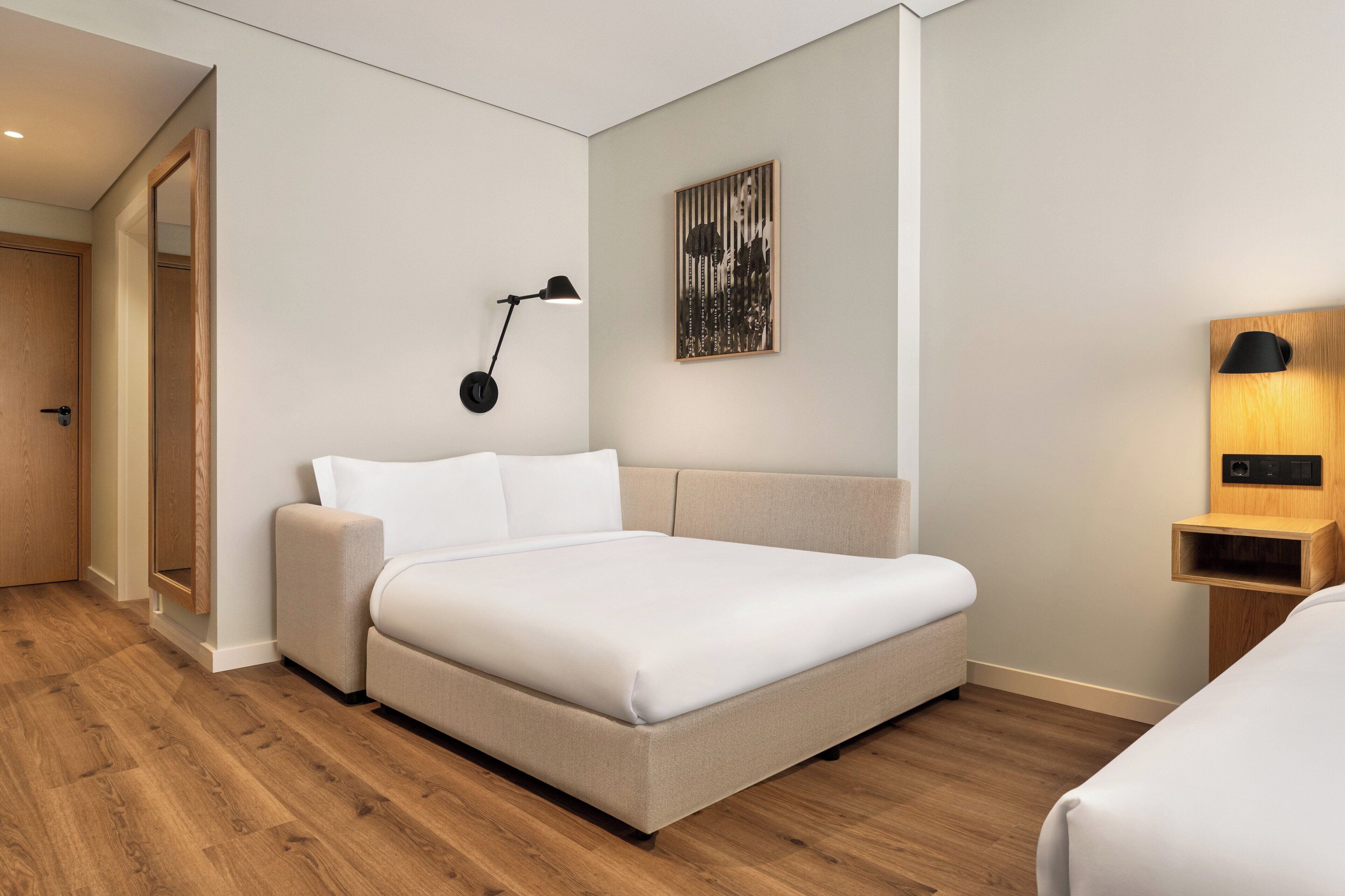 A modern hotel room with a large bed, a bedside table, a wall-mounted lamp, and a framed artwork.