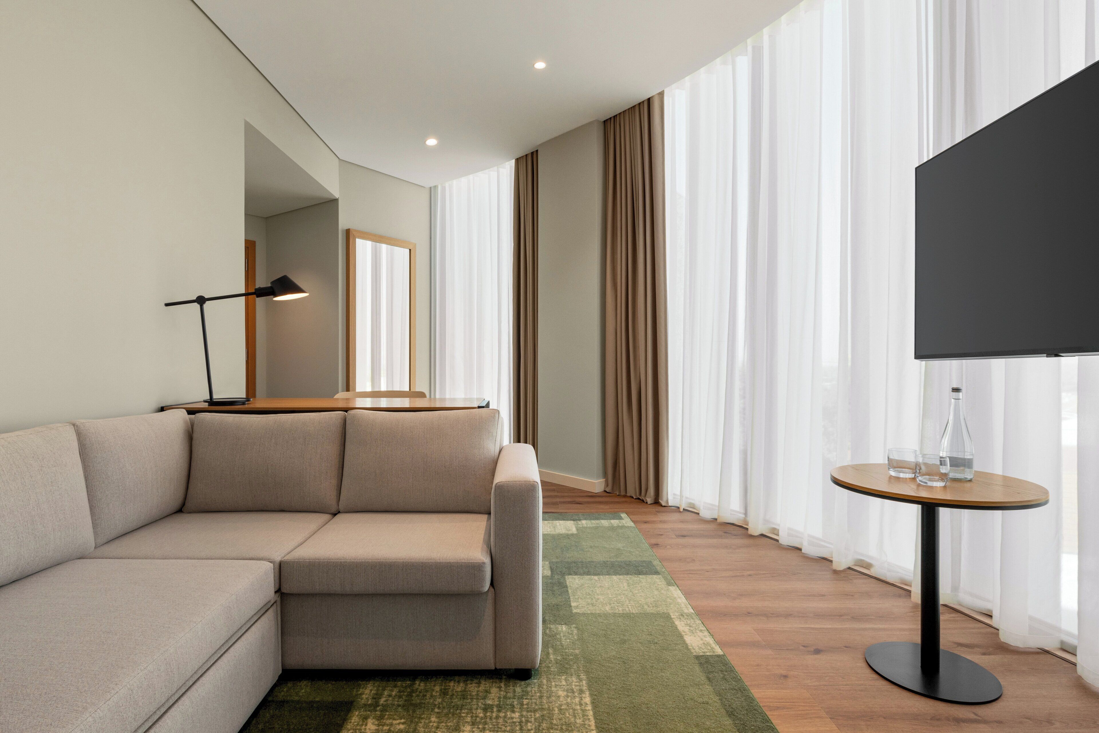 A modern living room with a sectional sofa, a round table with a bottle and glasses, a flat-screen TV, and a large window with sheer curtains.