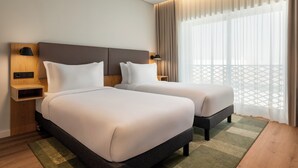 A hotel room with two beds, a wooden nightstand, a wall-mounted lamp, and a view of a balcony with a lattice structure. - Holiday Inn Beja by IHG (Beja)