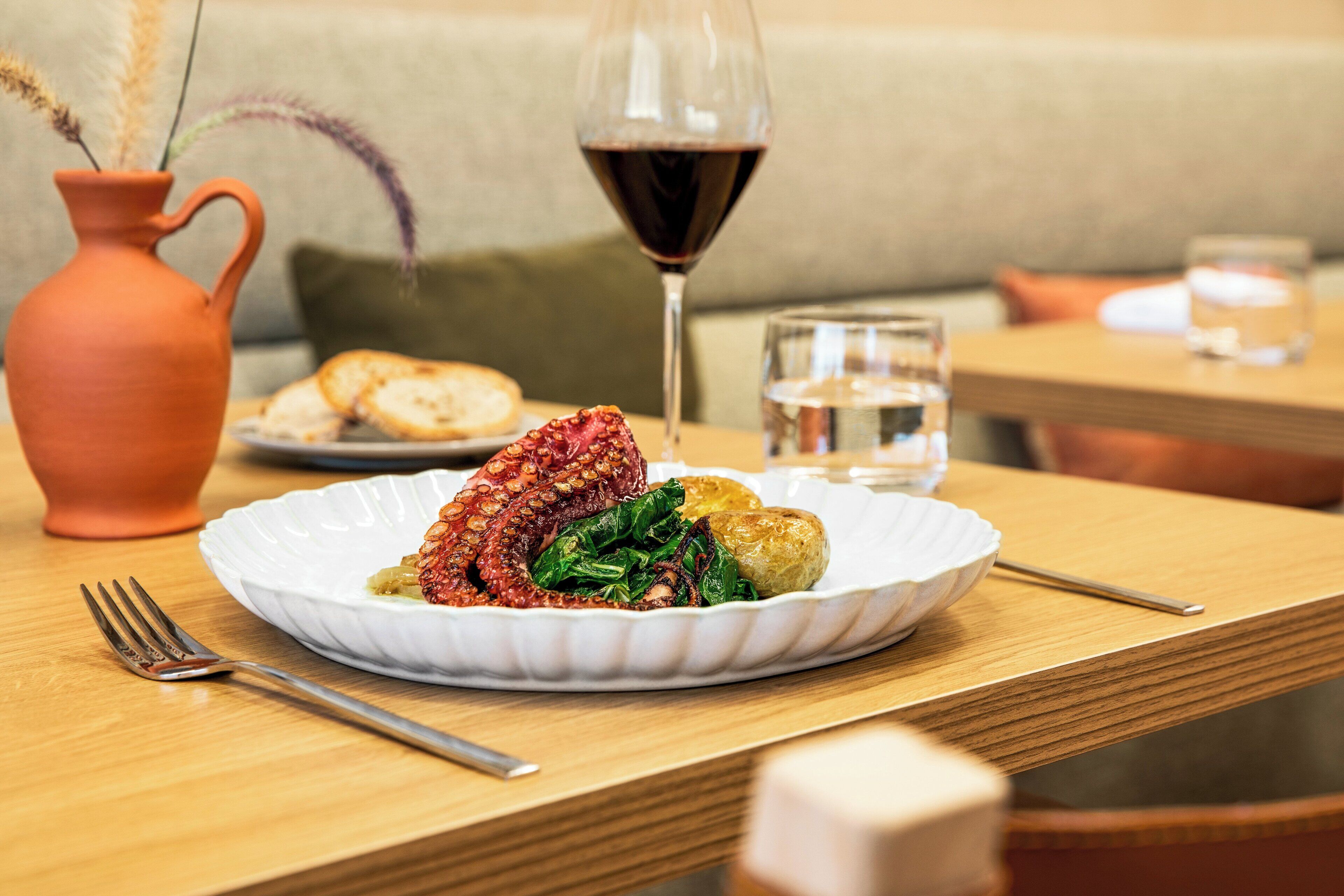 Una mesa de comedor con un plato de comida, una copa de vino tinto y un florero con plantas secas.