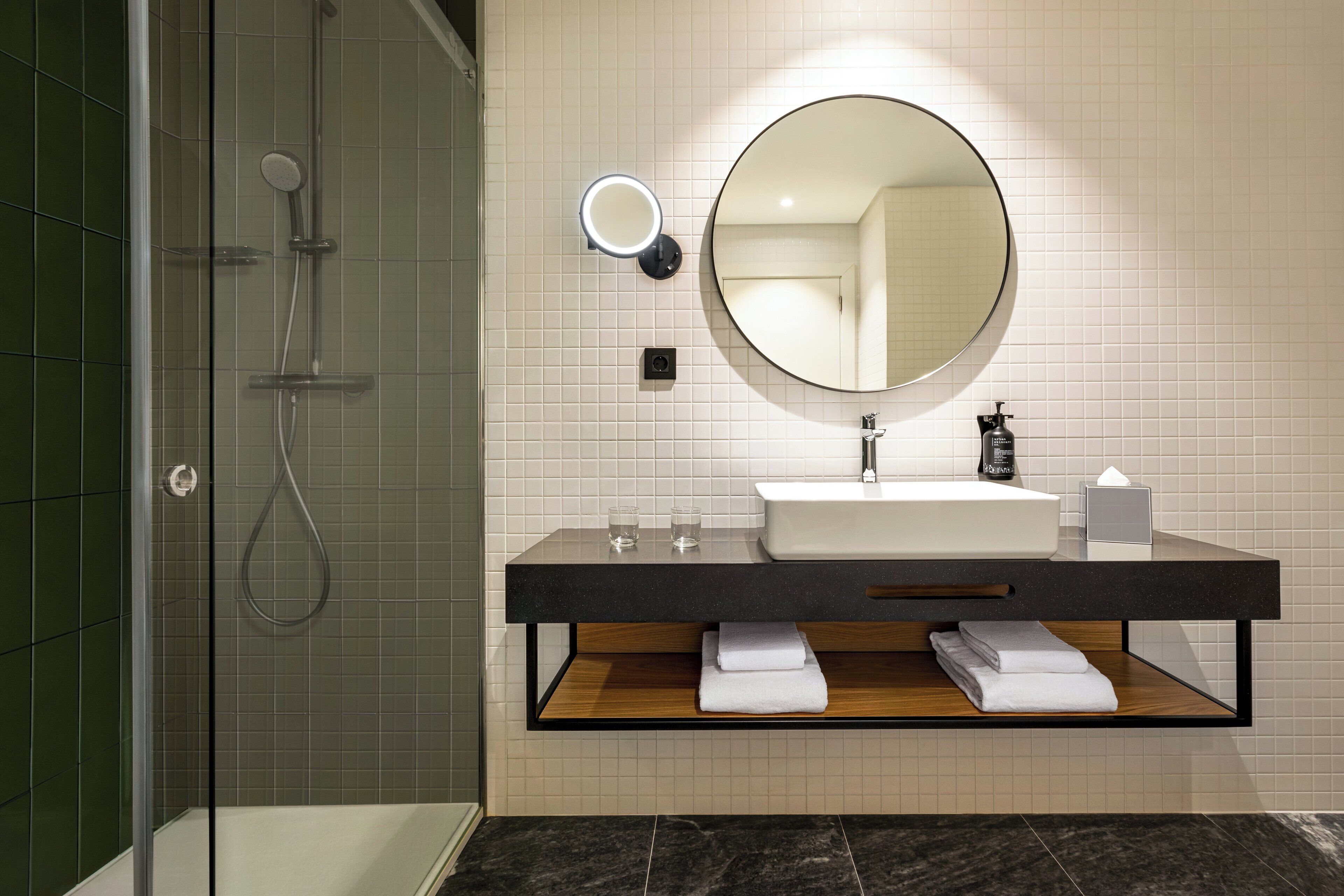 A modern bathroom with a glass-enclosed shower, a large round mirror, a dark countertop, and neatly folded towels.