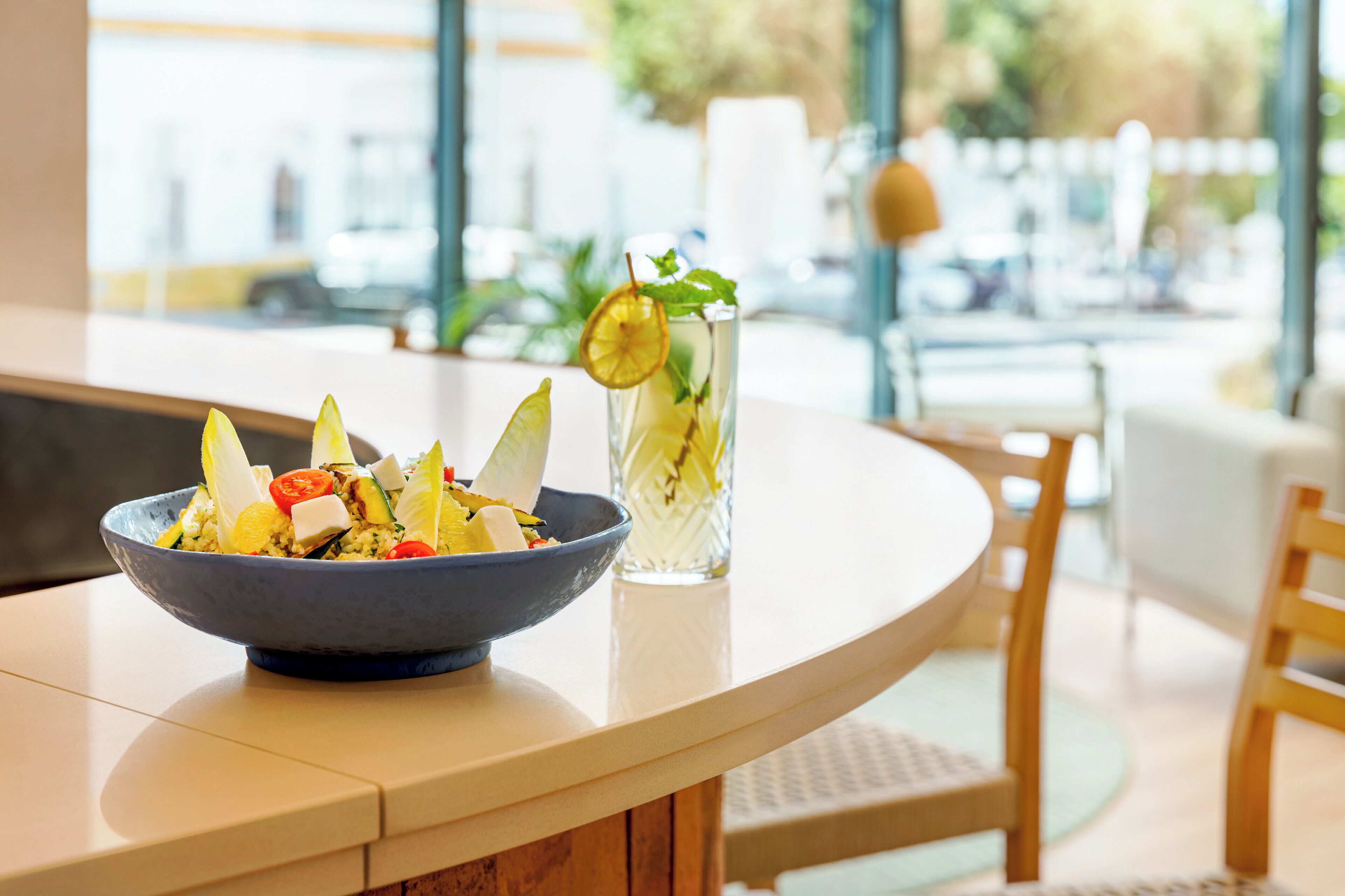 Une table à manger avec un saladier rempli de salade et un grand verre d’eau contenant une tranche de citron et quelques feuilles de menthe.