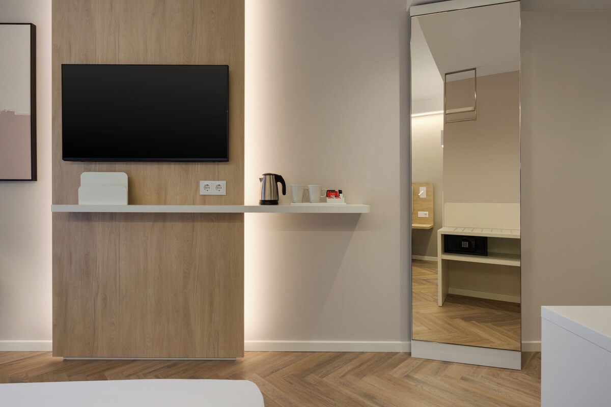 Una camera d'albergo moderna con una parete in legno decorativa, una TV a schermo piatto montata sopra un mobile bianco, una macchina per il caffè e uno specchio che riflette l'ambiente.