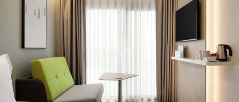 A hotel room with a bed, a green armchair, a small table, a flat-screen TV, and a coffee machine on a shelf.