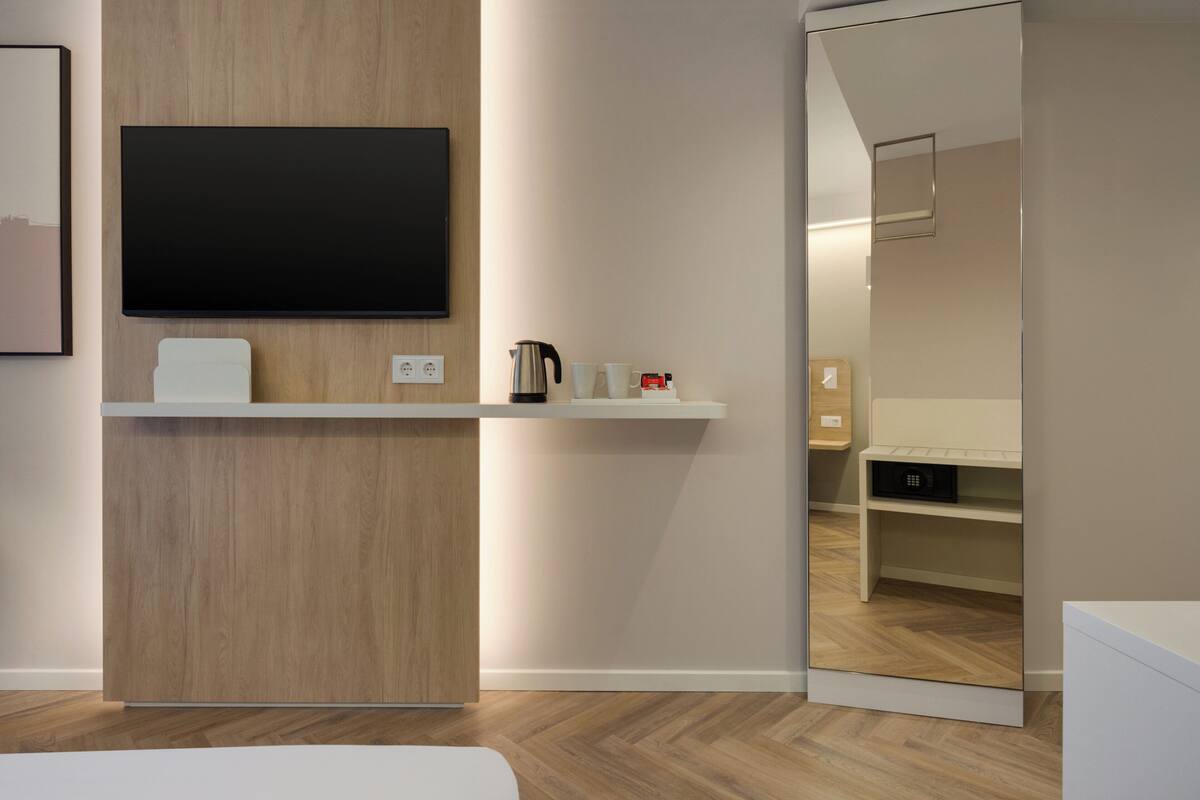 A modern hotel room with a wooden accent wall, a flat-screen TV mounted above a white console, a coffee maker, and a mirror reflecting the room.