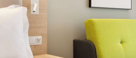 A hotel room with a bed, a nightstand, a lamp, and a wall-mounted power outlet.