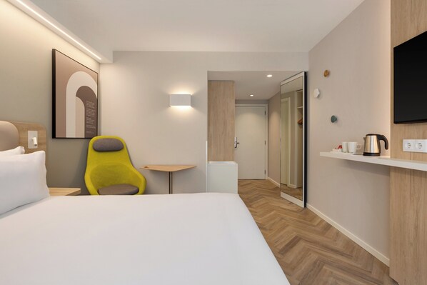 Una habitación de hotel con una cama, una silla amarilla, una mesita pequeña, un televisor montado en la pared y una cafetera en un estante.