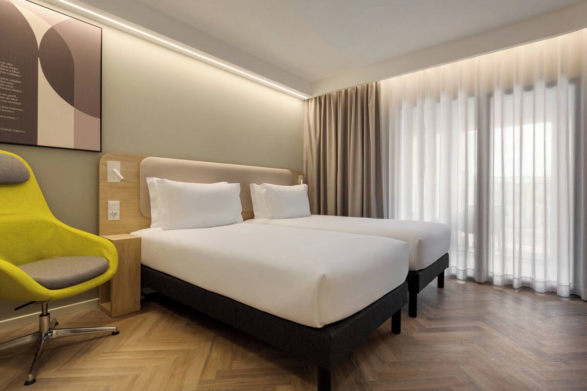 A hotel room with a large bed, a bedside table, a chair, and a window with curtains.