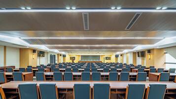 Uma sala de conferência com fileiras de mesas e cadeiras, iluminação no teto e sistema de ventilação.