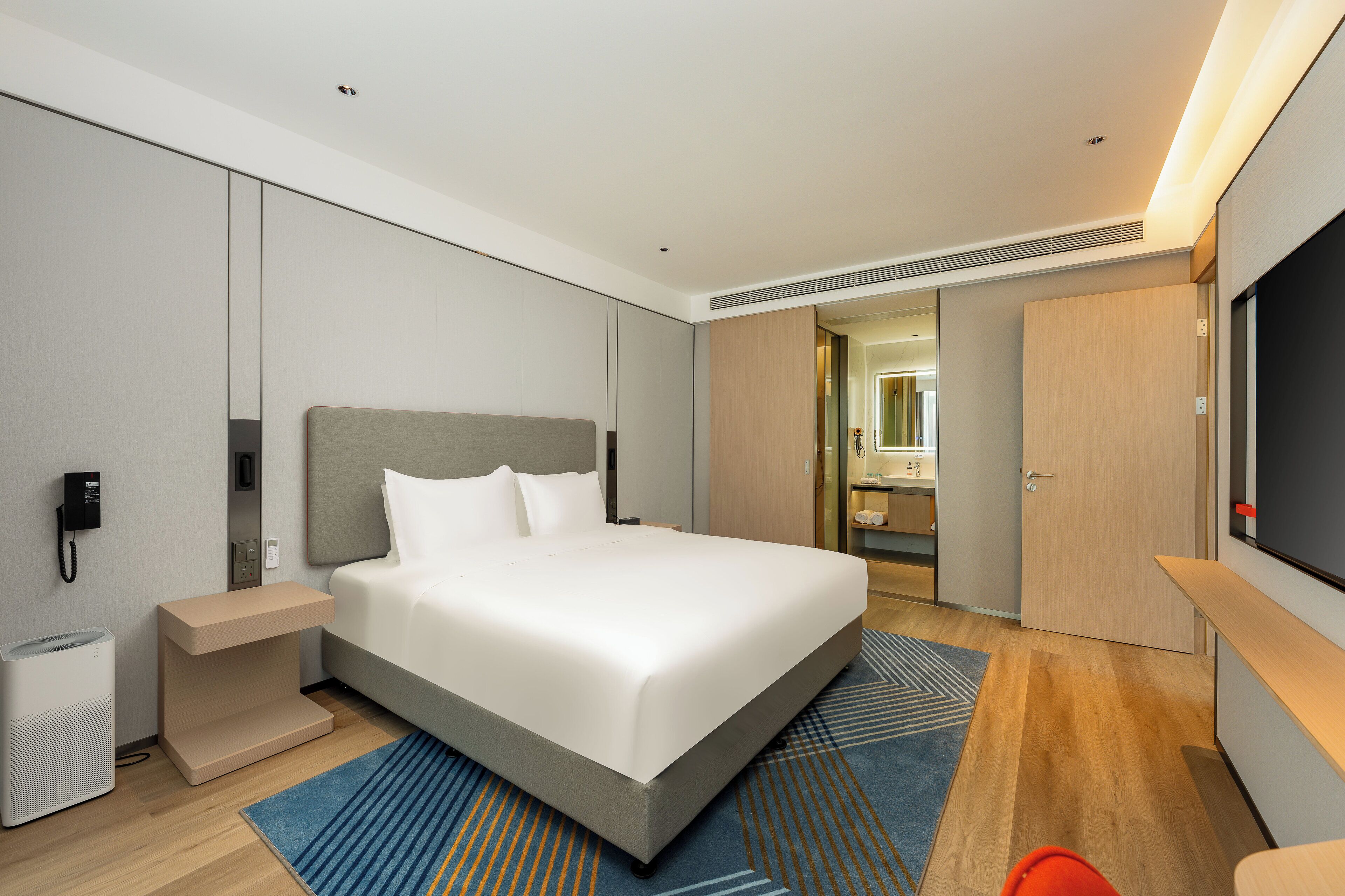 A modern hotel room with a large bed, a bedside table, a TV, and a bathroom visible through an open door.