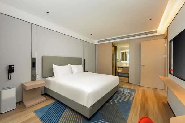 A modern hotel room with a large bed, a bedside table, a TV, and a bathroom visible through an open door.