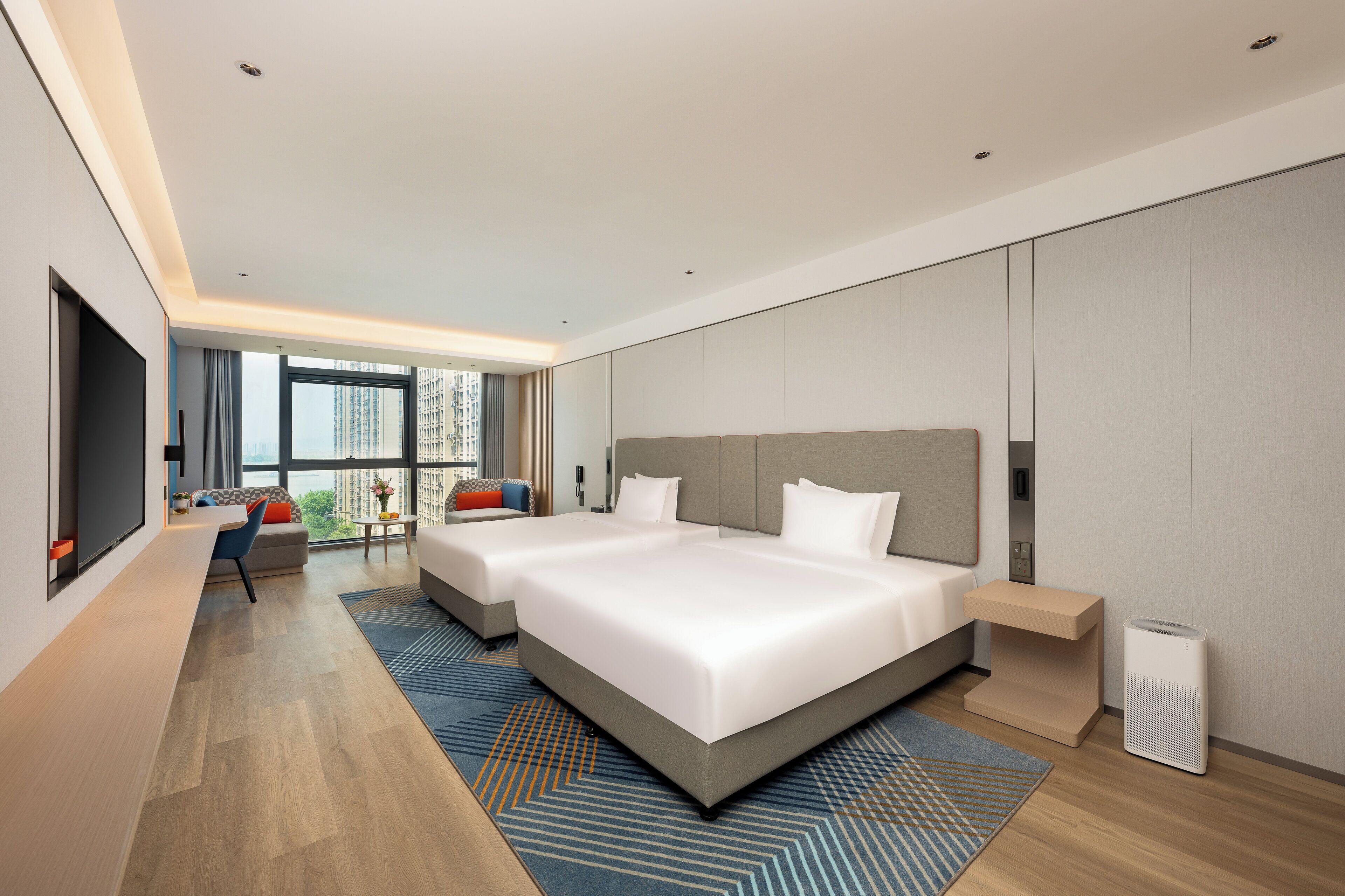 A modern hotel room with two large beds, a flat-screen TV, a small seating area, and a view of the city.