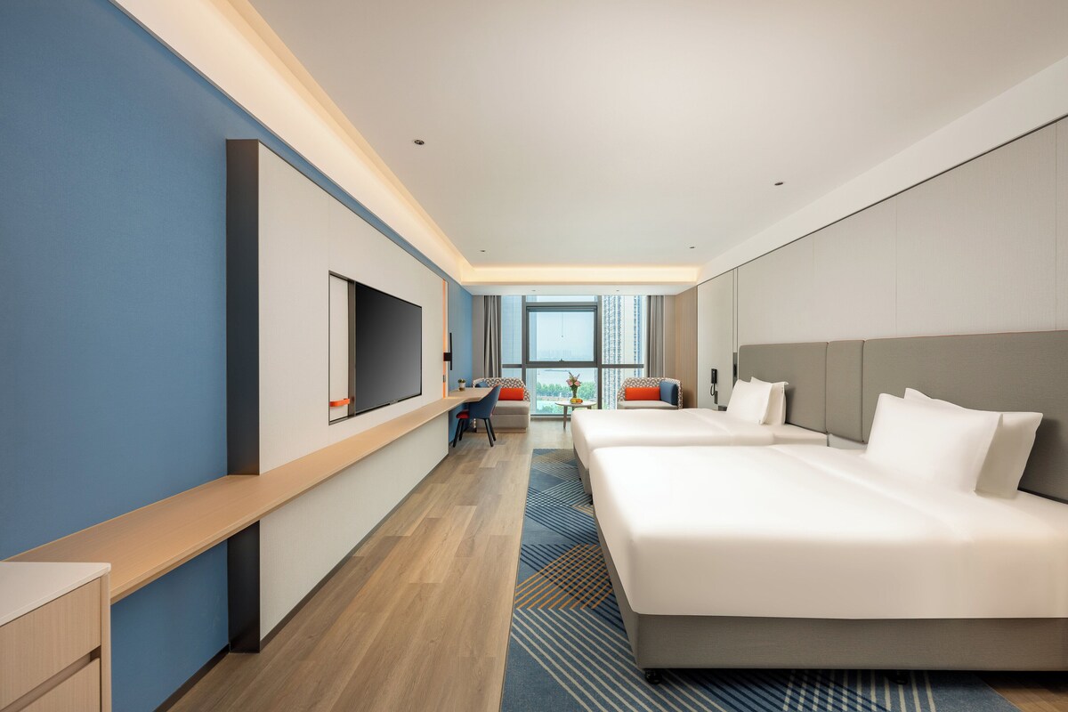 A modern hotel room with a large bed, a desk with a chair, a television, and a window with a city view.
