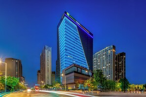 A high-rise building with a prominent illuminated facade, surrounded by other tall buildings and a busy street with moving vehicles.