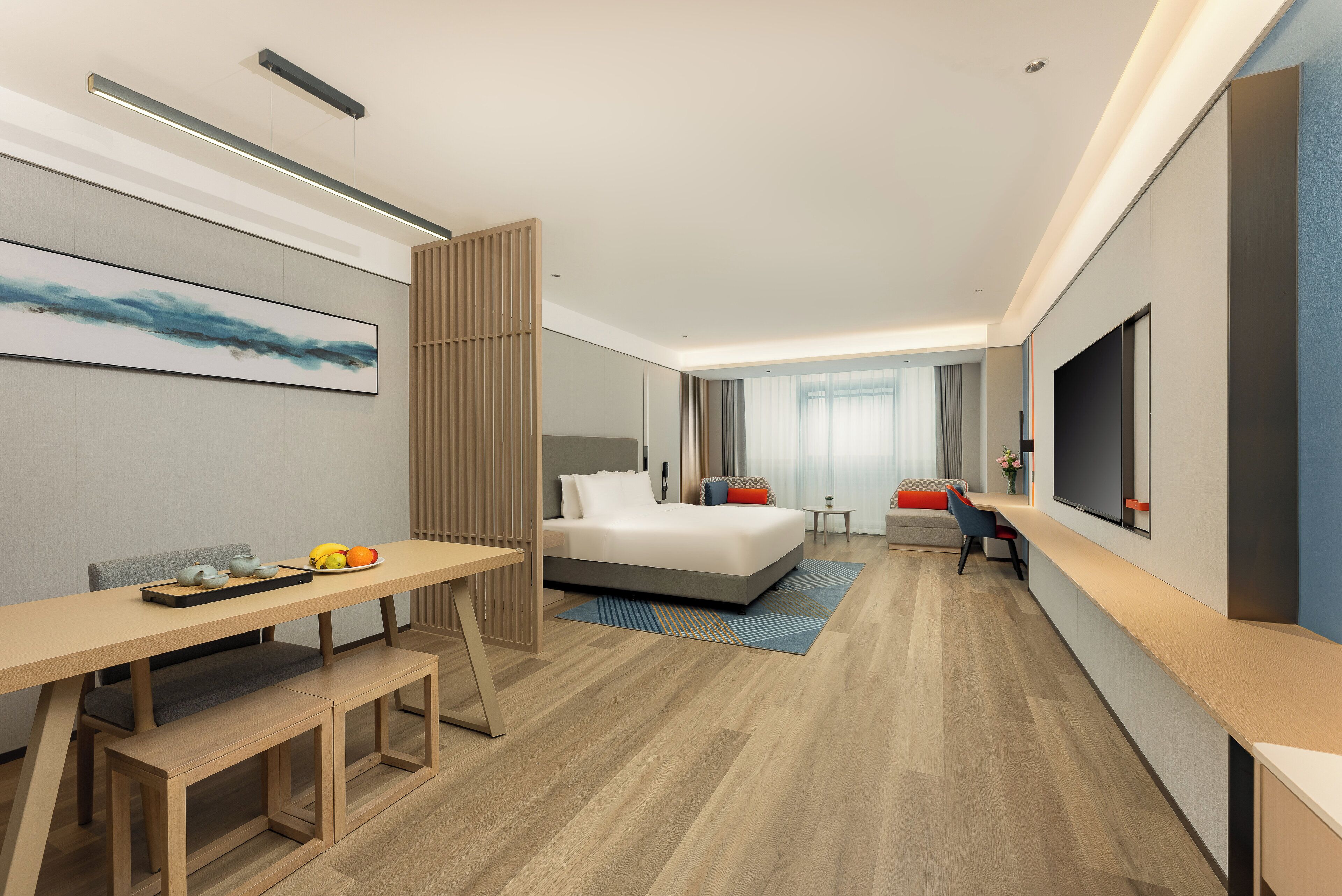 A modern hotel room with a bed, bedside table, desk, and a large TV.