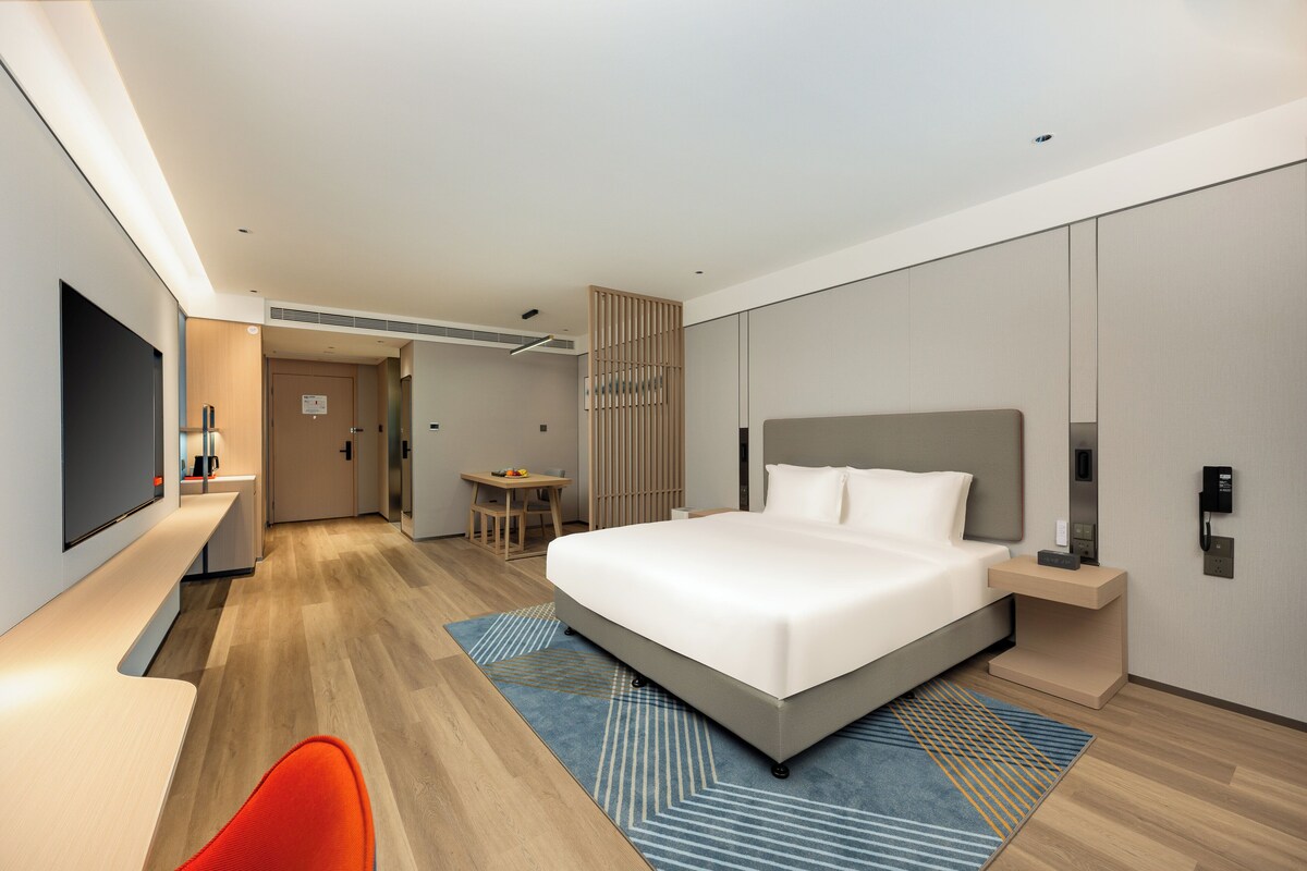 A modern hotel room with a large bed, a desk, a TV, and a seating area.