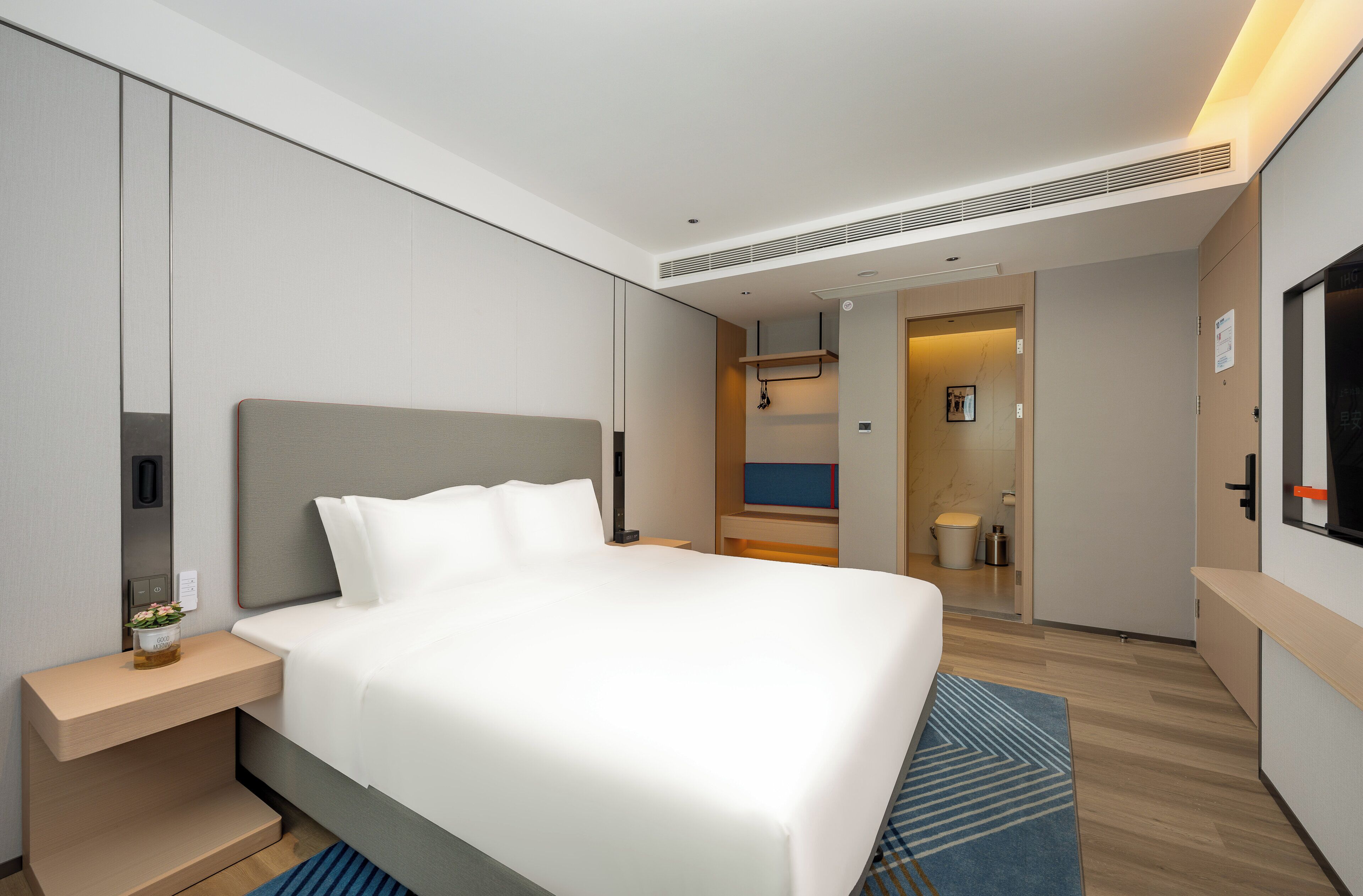 A modern hotel room with a large bed, bedside table, and a small bedside table with a vase.